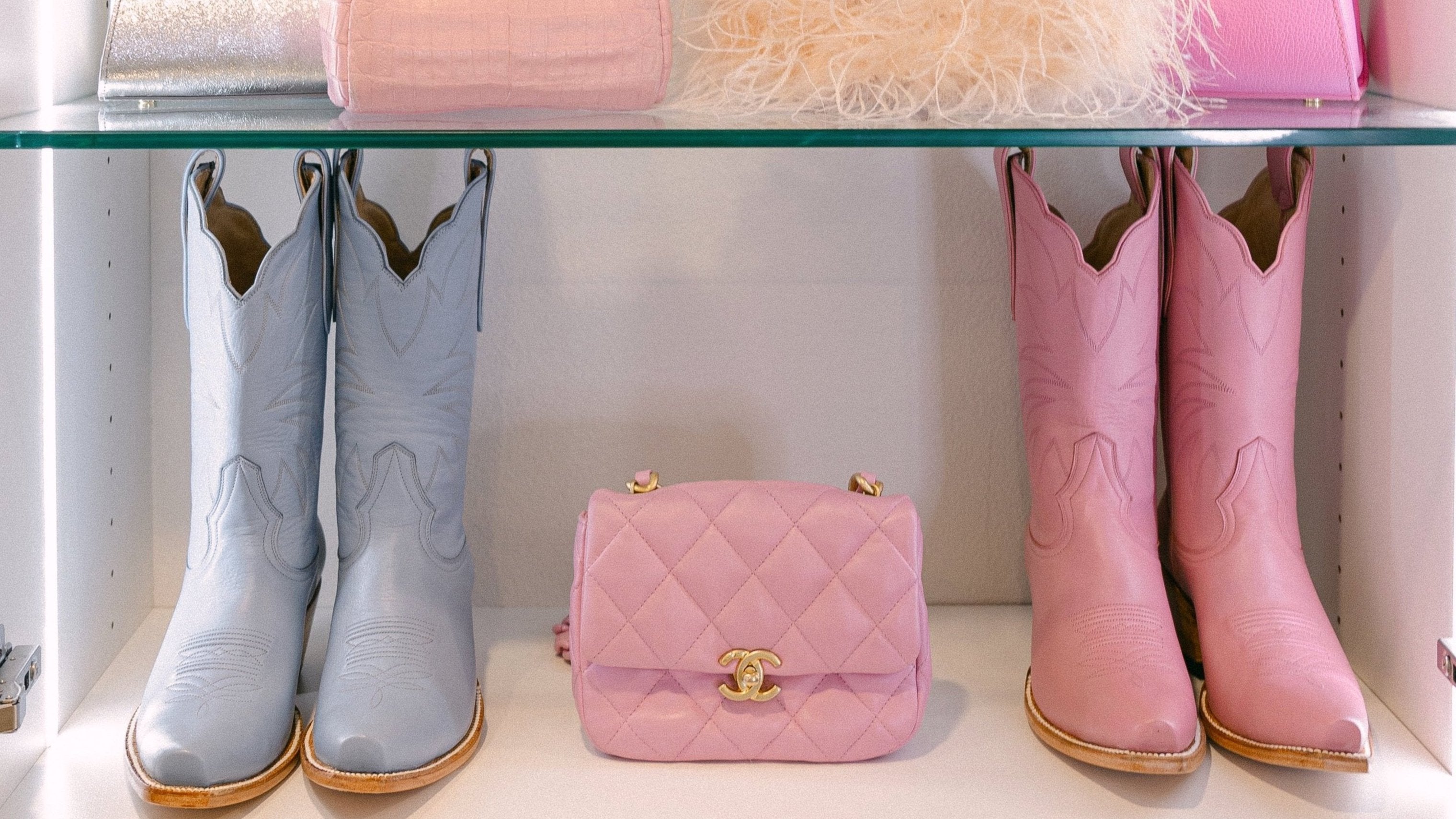 Two pairs of cowgirl cowboy boots, one powder baby blue and one light petal pink, on a shelf with a pink chanel handbag.