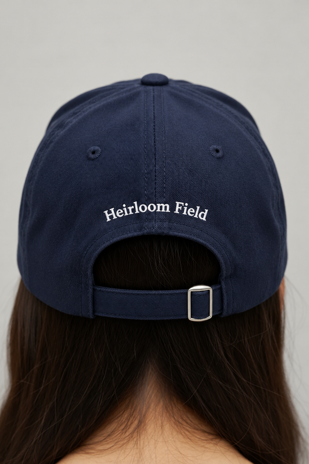 Back view of a navy cap worn over long hair, with Heirloom Field embroidery and a metal strap buckle.