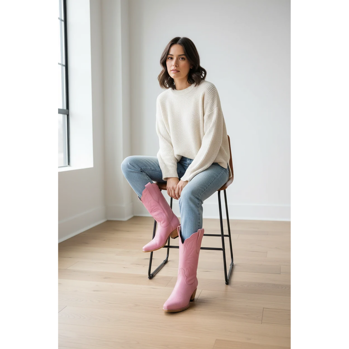 Woman seated on a stool wearing pink midi boots with jeans and a cream sweater.