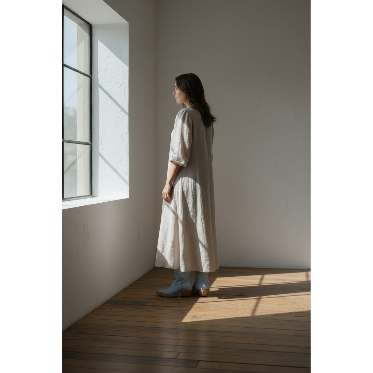 Woman wearing pale-blue boots with a light midi dress, standing beside a window.