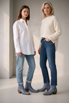 Two women mother daughter wearing white shirts and blue jeans with gray boots against a neutral background