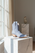 Blue cowboy boots on a white pedestal in a bright room with a vase in the background.