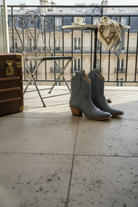 Pair of gray blue alligator cowgirl boots on a balcony with a suitcase and table in the background.