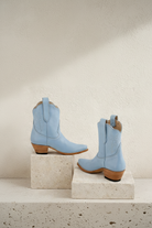 Pair of light blue cowboy boots with brown soles on a beige stone surface. lily of the valley embroidery