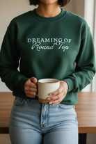 Person wearing a green sweatshirt with 'Dreaming of Round Top' text, holding a mug.