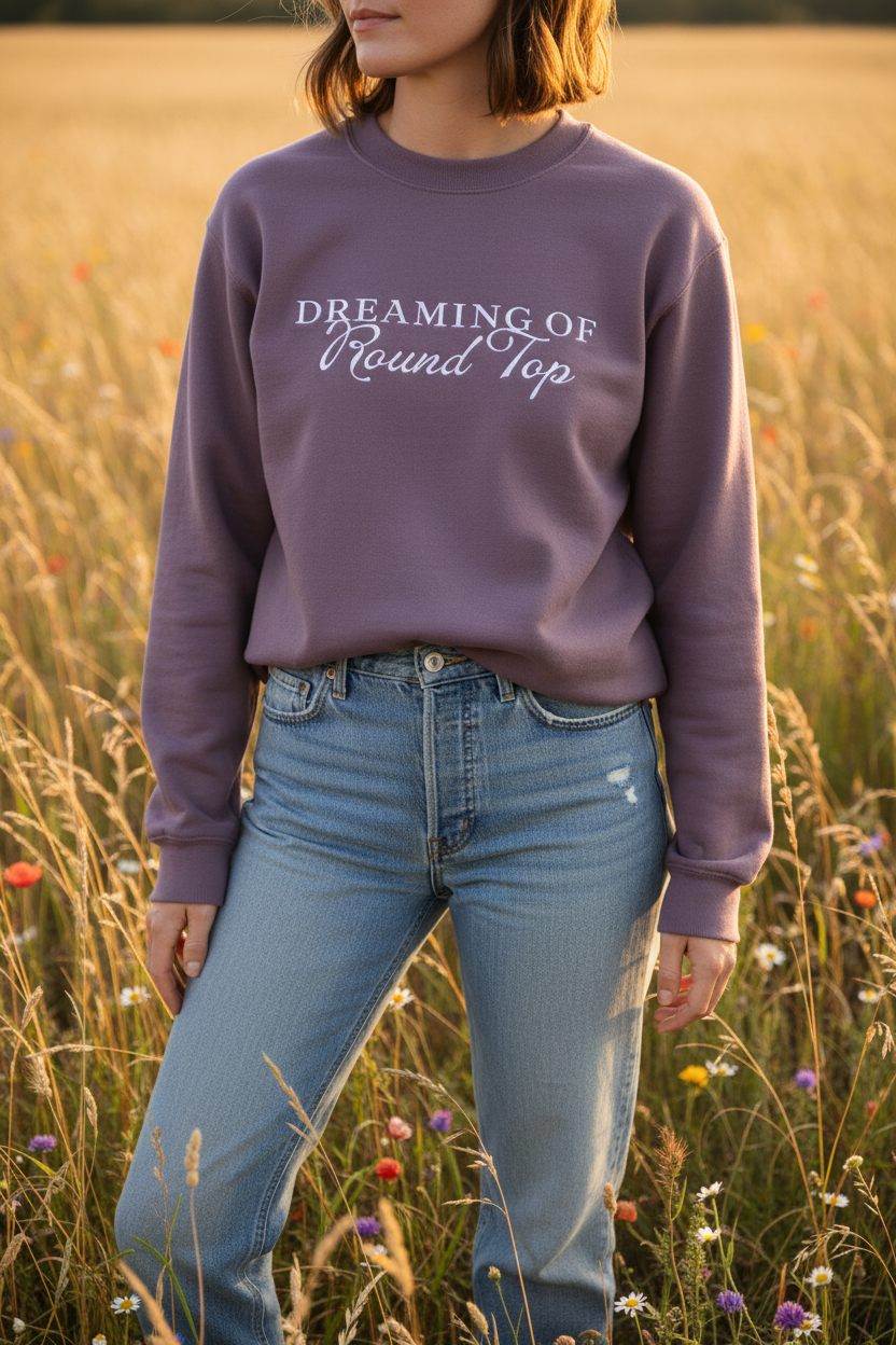 Person wearing a purple sweatshirt with text Dreaming of Round Top embroidered standing in a field of tall grass and wildflowers.