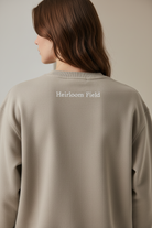 Person wearing a beige sweatshirt with 'Heirloom Field' text on the back against a neutral background