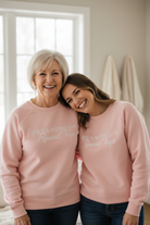 Two women wearing matching pink Dreaming of Round Top sweatshirts with text, standing close together indoors.
