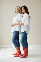 Two women wearing white tops and blue jeans with  luxury handcrafted deep red boots standing close together against a plain background.