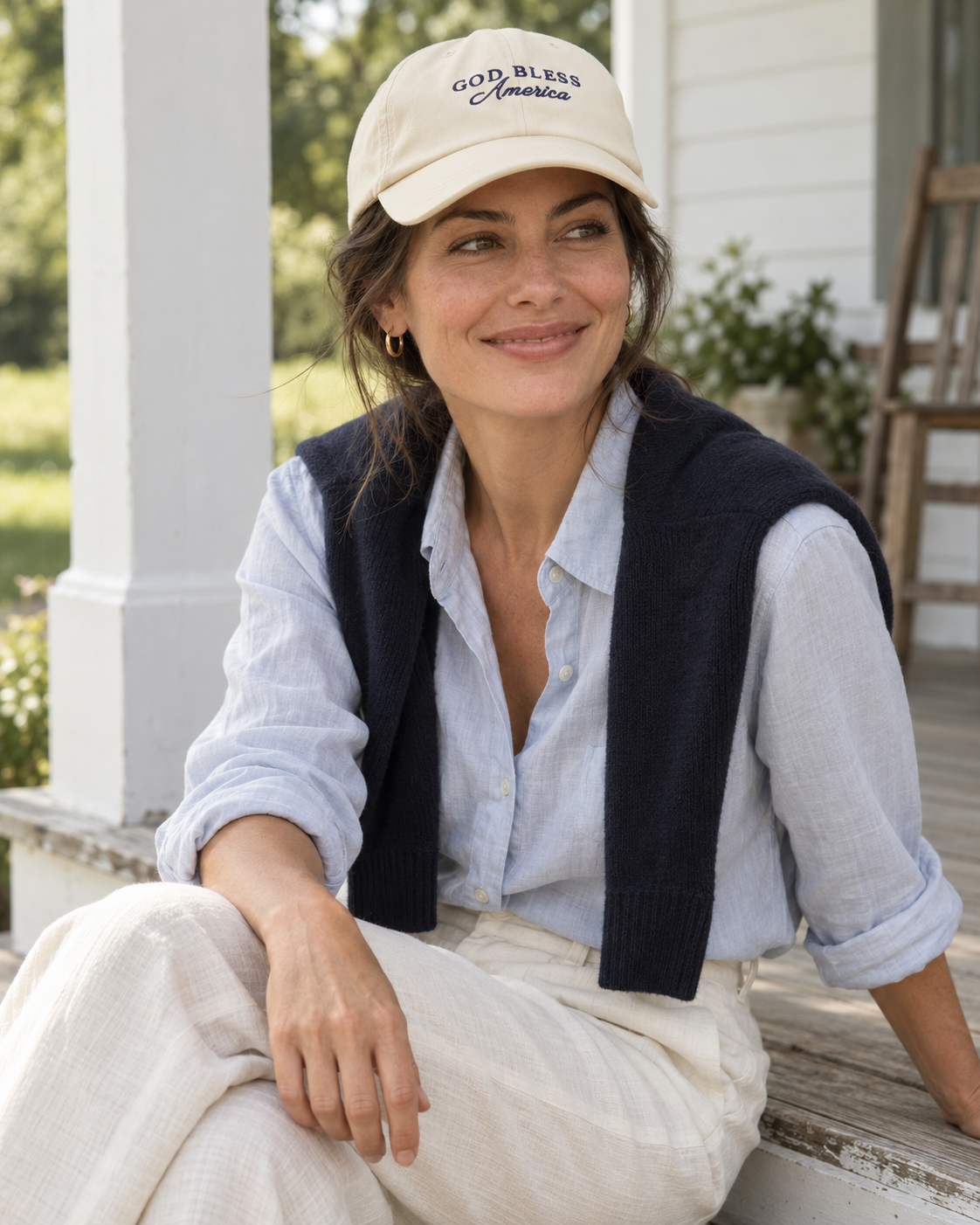 Woman wearing the cream God Bless America cap with a pale blue shirt and plain navy sweater on a white porch.