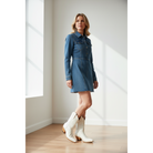 Woman wearing white midi boots with a dark denim dress by a window.