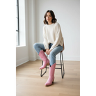 Woman seated on a stool wearing pink midi boots with jeans and a cream sweater.