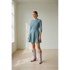 Woman wearing pale-lavender midi boots with a blue dress in a sunlit room.