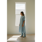 Woman in a blue midi dress wearing pale-blue boots beside a tall window.