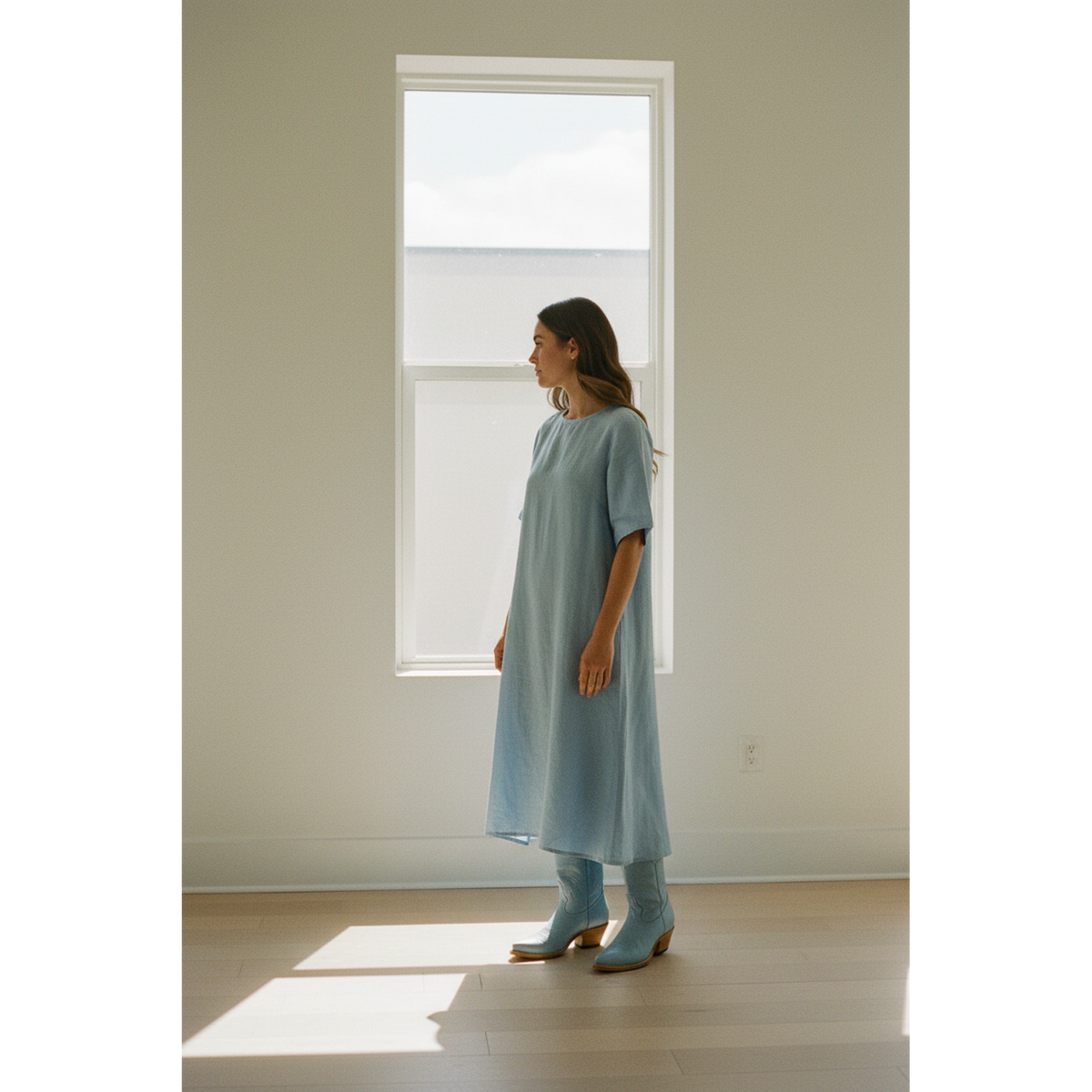 Woman in a blue midi dress wearing pale-blue boots beside a tall window.