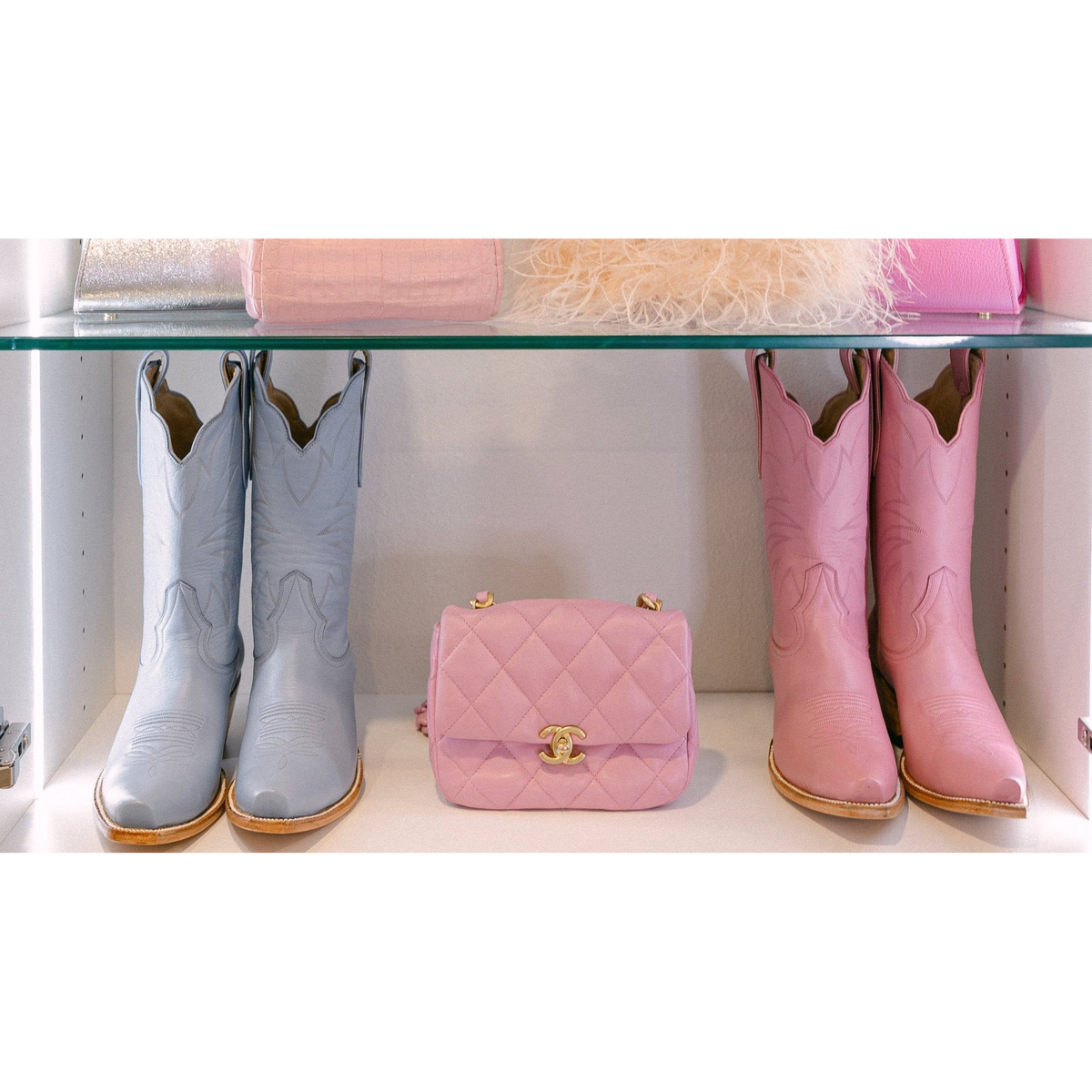 Two pairs of cowgirl cowboy boots, one powder baby blue and one light petal pink, on a shelf with a pink chanel handbag.